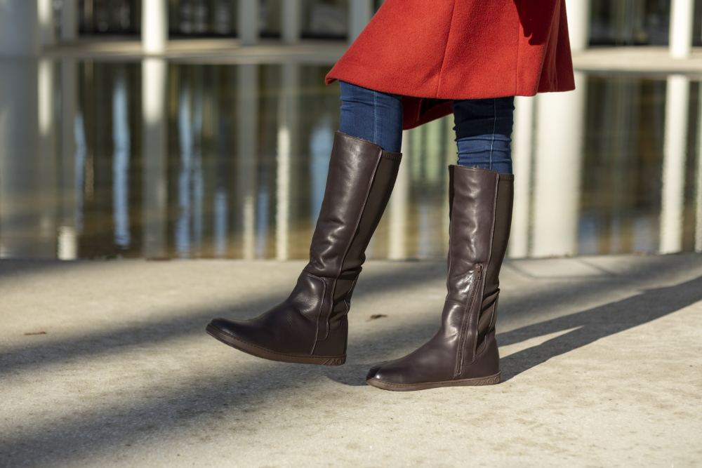 Eine Person in rotem Mantel und dunkelblauer Jeans spaziert in hohen, dunkelbraunen ZAQQ QATCH Winter Brown-Stiefeln aus edlem Nappaleder durch die Natur. Im Hintergrund spiegeln sich Säulen im Wasser, der Boden ist sonnenbeschienen.