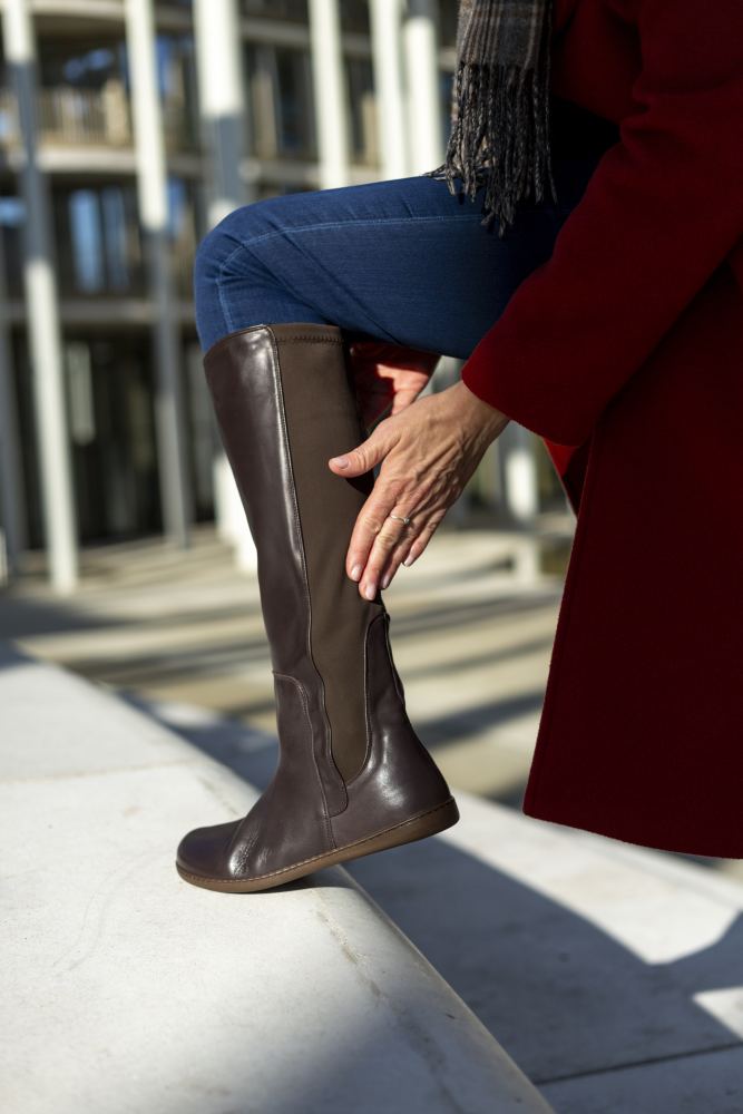 Jemand trägt einen roten Mantel und blaue Jeans und rückt seinen ZAQQ QATCH Nappa Brown-Stiefel zurecht und stellt ihn auf einen Sims. Hinter ihnen hebt ein modernes Gebäude mit großen Fenstern die ideale Kombination aus urbanem Stil und Funktionalität hervor.