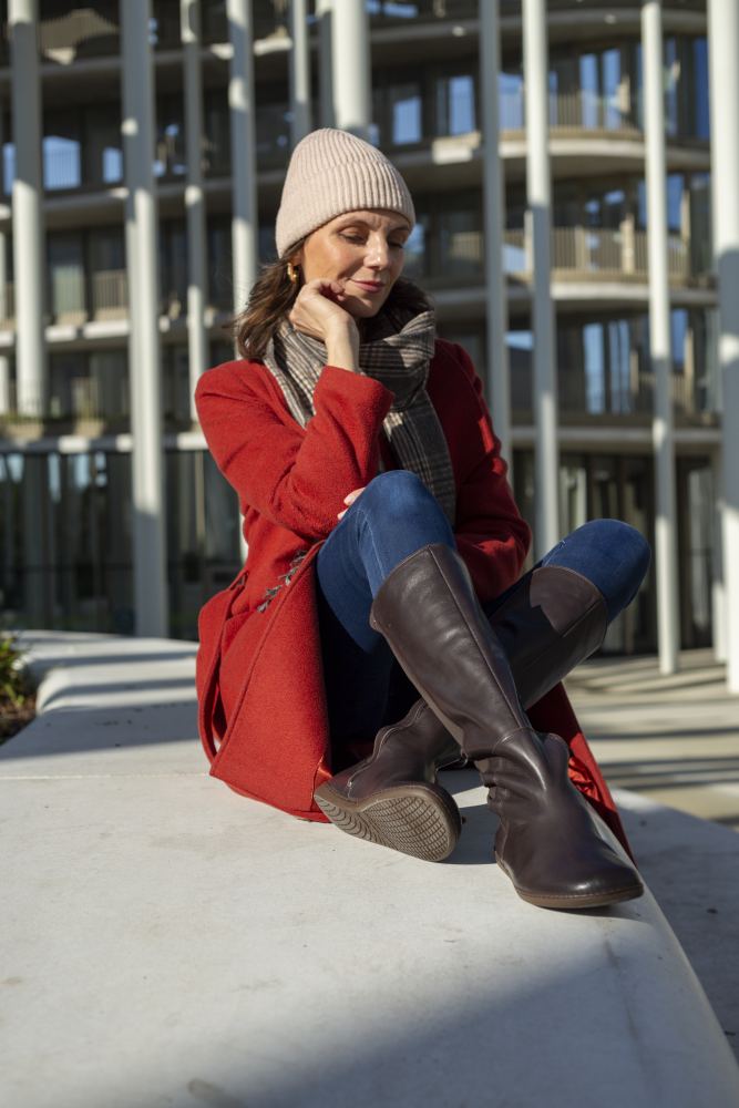 Eine Person in rotem Mantel und Jeans, die elegante ZAQQ QATCH Nappa Brown-Stiefel trägt, sitzt nachdenklich auf einem Betonsims. Sie trägt eine beige Strickmütze und einen karierten Schal. Im Hintergrund steht ein modernes, mit Säulen geschmücktes Gebäude.