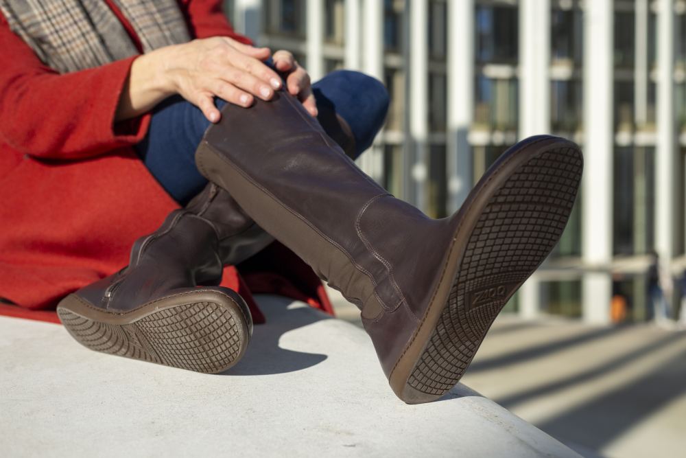 Eine Person in einem roten Mantel, blauen Jeans und kniehohen Stiefeln QATCH Nappa Brown von ZAQQ sitzt draußen und hat ihre Füße auf einer Oberfläche. Im Hintergrund ist ein modernes Gebäude mit vertikalen weißen Linien zu sehen.