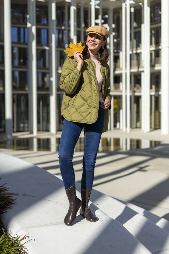 Eine lächelnde Frau steht im Freien und hält gelbe Herbstblätter in der Hand. Sie trägt eine grüne Steppjacke, blaue Jeans und braune ATTRAQT Stretch Nappa Boots von ZAQQ aus Ziegennappaleder. Eine beigefarbene Mütze rundet ihren Look ab, während sie vor der Kulisse eines modernen Glasgebäudes posiert.