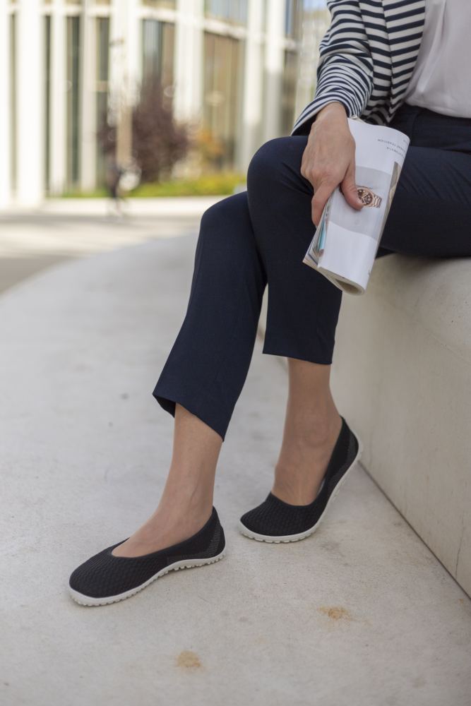 Eine Person in eleganten SUQAR Black Slippern von ZAQQ und marineblauen Hosen sitzt auf einem Betonsims und hält eine zusammengerollte Zeitschrift in der Hand. Das stilvolle Schuhwerk verleiht einen subtilen, femininen Look. Im Hintergrund ist ein modernes Gebäude mit hohen Fenstern zu sehen.