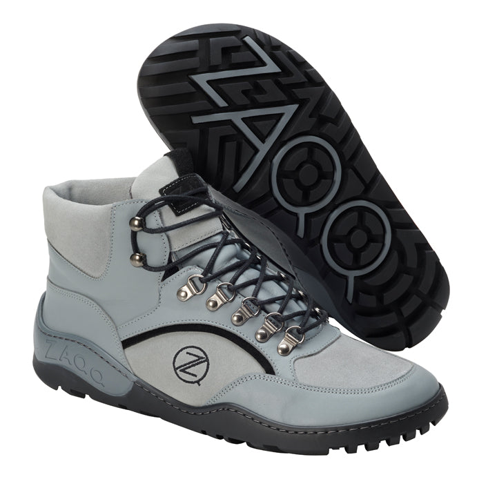 Eine Nahaufnahme der TREQ Grey Waterproof Wanderstiefel von ZAQQ, die ihr stilvolles graues Erscheinungsbild mit schwarzen Sohlen zeigt. Die Stiefel sind mit Metallösen und schwarzen Schnürsenkeln ausgestattet und tragen das ZAQQ-Logo sowohl an der Seite als auch an der Sohle. Ein Stiefel steht aufrecht, während der andere geneigt ist, um sein Sohlenmuster zu zeigen.