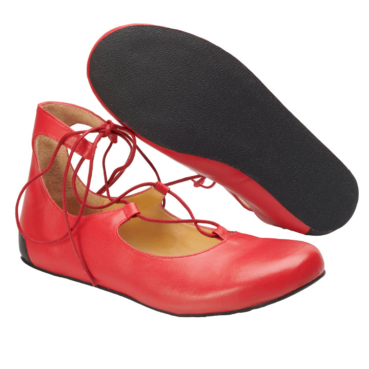 Ein Paar QISS Red Ballerinas von ZAQQ, gefertigt aus Premium Nappaleder in leuchtendem Rot, präsentiert schwarze Sohlen und zarte Schnürriemen, die sich elegant um die Knöchel wickeln. Ein Schuh wird aufrecht präsentiert, während der andere geneigt ist, um seine Sohle hervorzuheben, die das Barfußgefühl verbessern soll.