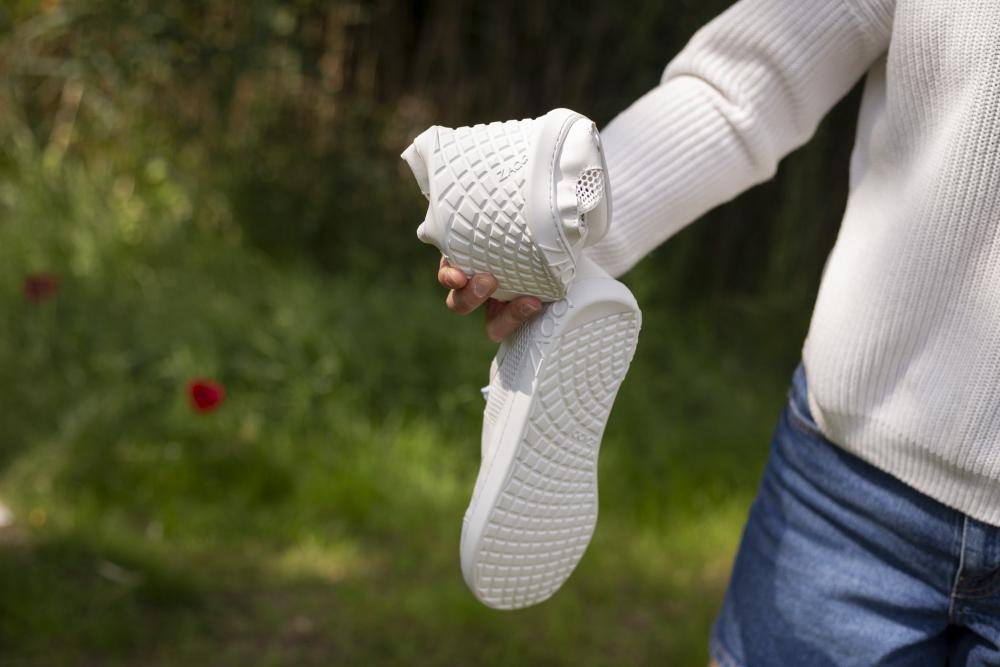 Auf einer Rasenfläche mit verschwommenem Laub im Hintergrund steht eine Person in einem weißen Pullover und blauen Jeans und hält ein Paar elegante weiße ALIQANTE-Sneaker von ZAQQ in der Hand, die für ihre strukturierte Sohle bekannt sind, die Komfort und Atmungsaktivität betont.