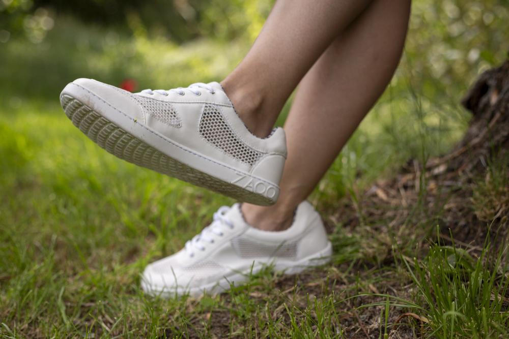 Eine Person, die den ALIQANTE White Sneaker von ZAQQ mit Mesh-Einsätzen und Nappaleder trägt, steht mit einem Fuß leicht angehoben auf Gras. Das üppige grüne Laub und ein Baumstamm im Hintergrund verstärken das Gefühl von Atmungsaktivität.