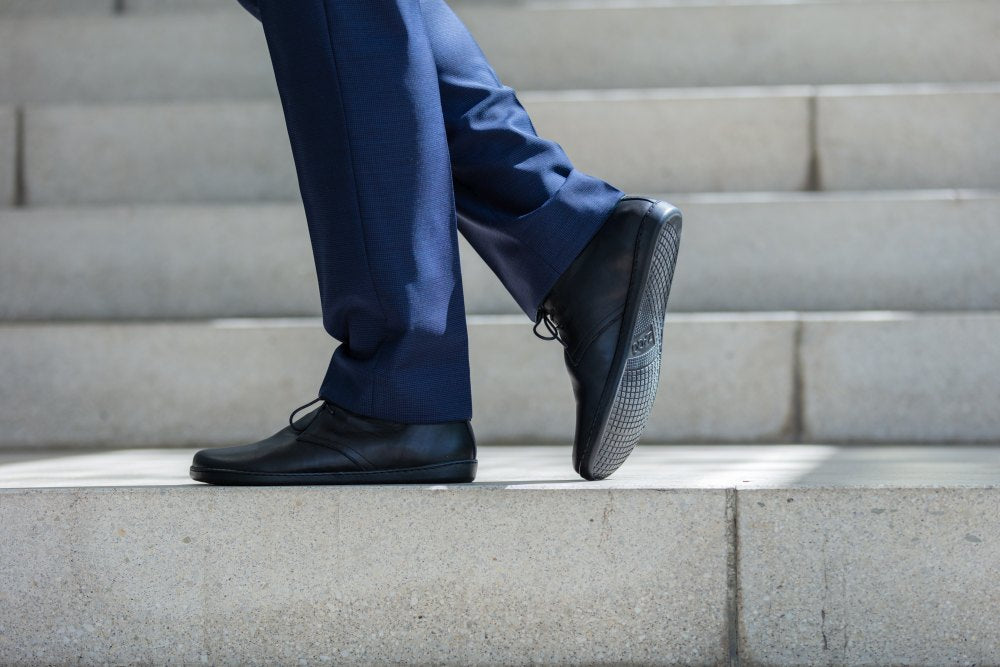 Eine Person in blauer Hose und schwarzen ARQ-Stiefeln von ZAQQ aus elegantem Glattleder geht eine Steintreppe hinauf (gezeigt von den Knien abwärts). Die Treppe befindet sich in einer gut beleuchteten Umgebung im Freien.