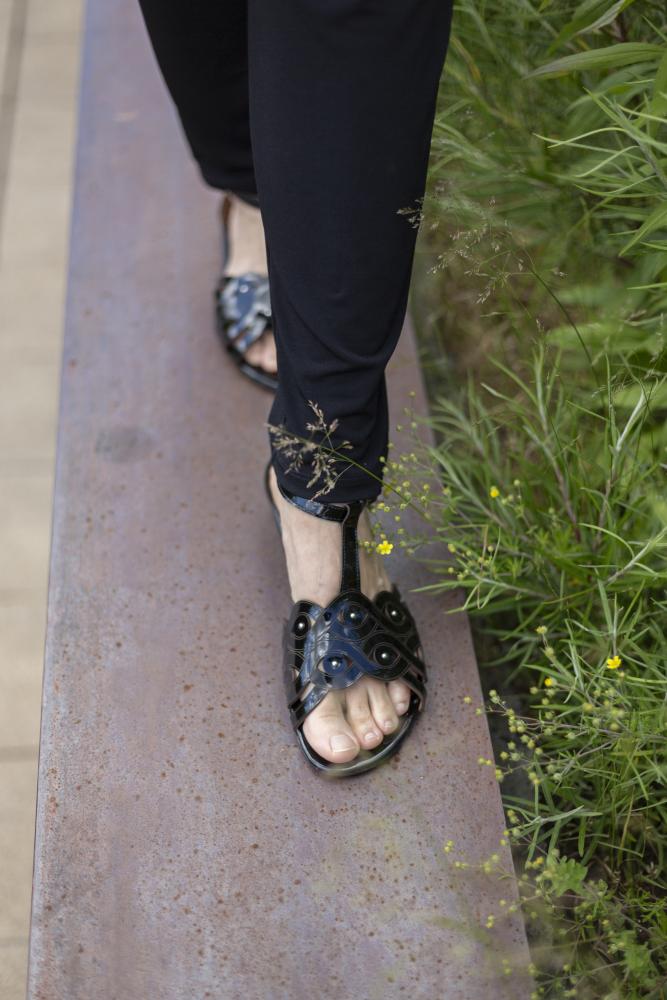 Eine Person in den Sandalen BOUQET Black von ZAQQ und einer schwarzen Hose läuft auf einem schmalen Metallträger. Neben dem Träger wachsen grünes Gras und kleine gelbe Blumen, die bei jedem Schritt die Rutschfestigkeit der Sandalen unterstreichen.