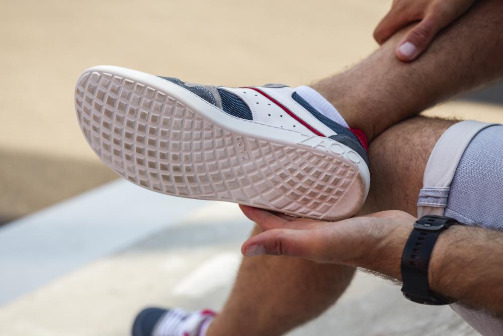 Eine Person sitzt draußen in einem Paar BREAQ Darkblue-Sneakers von ZAQQ und zeigt das Profilmuster mit der weißen Sohle. Sie hält ihren Knöchel und enthüllt eine elegante Smartwatch an ihrem Handgelenk. Die Sneaker sind in einer Palette von dunkelblauen, grauen und roten Farbtönen erhältlich.