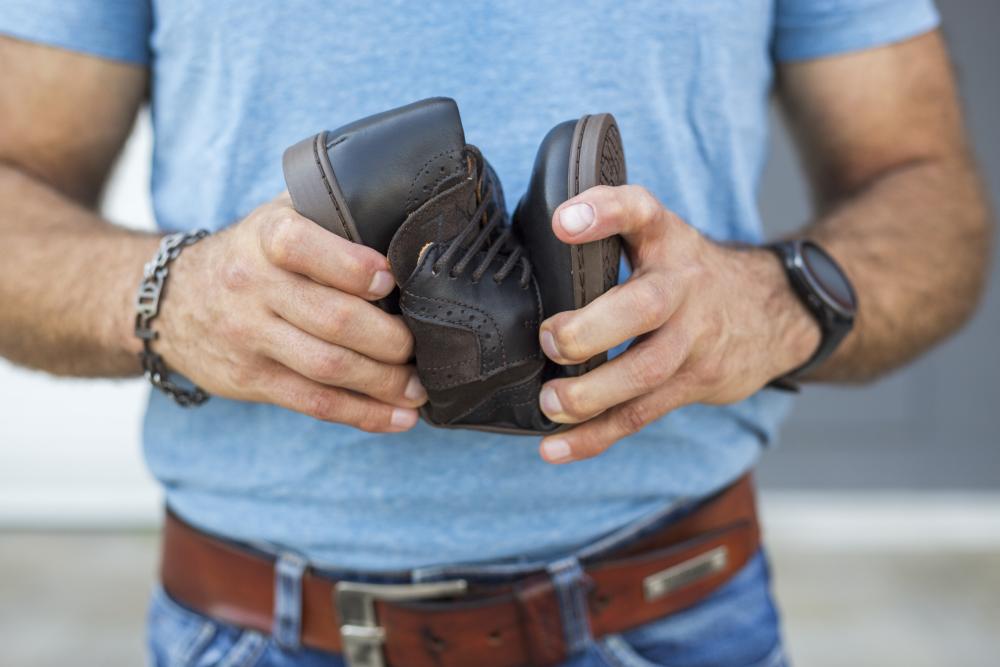 Eine Person in blauem Hemd und Jeans hält ein Paar kleine braune ZAQQ BRIQ Brogue Tonka Babyschuhe in der Hand, dazu eine Uhr und ein Metallarmband.