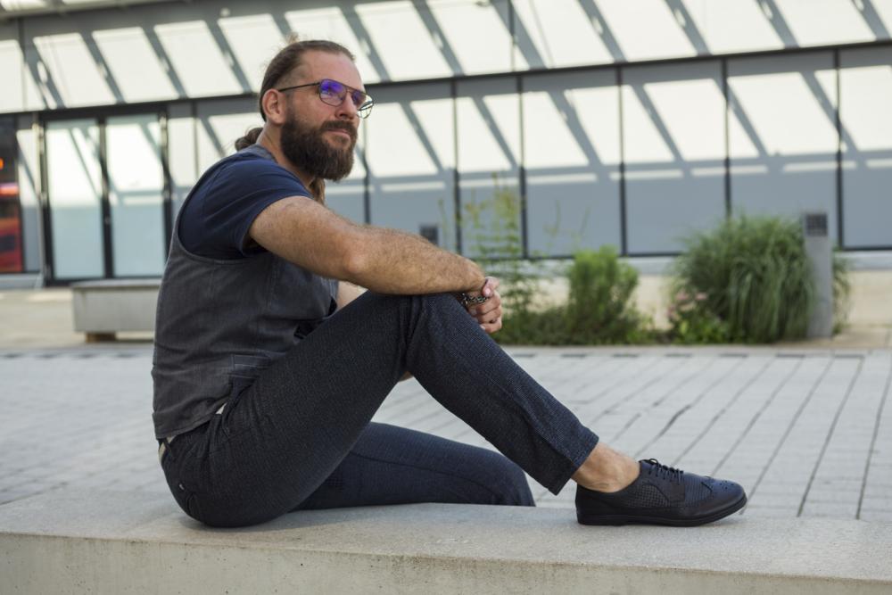 Ein Mann mit Bart, Brille, Weste und dunkler Hose, die zu seinen Business-Schuhen ZAQQ BRIQ Brogue Tresse Black passt, sitzt auf einer Betonbank in einer städtischen Umgebung. Er blickt nachdenklich in die Ferne, während Schatten einer Glasstruktur Muster auf den Boden werfen.