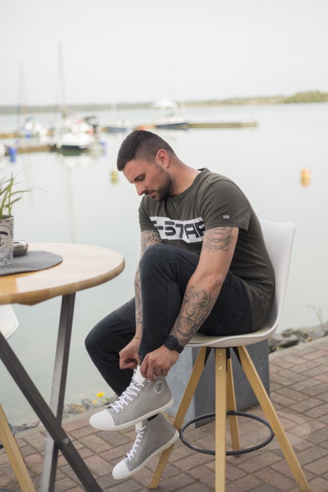 Ein Mann sitzt auf einem weißen Stuhl in Ufernähe und bindet die Schnürsenkel seiner grauen ZAQQ CHUQQS High-Top-Sneakers. Er trägt ein grünes T-Shirt und eine schwarze Hose, während im Hintergrund Boote treiben.