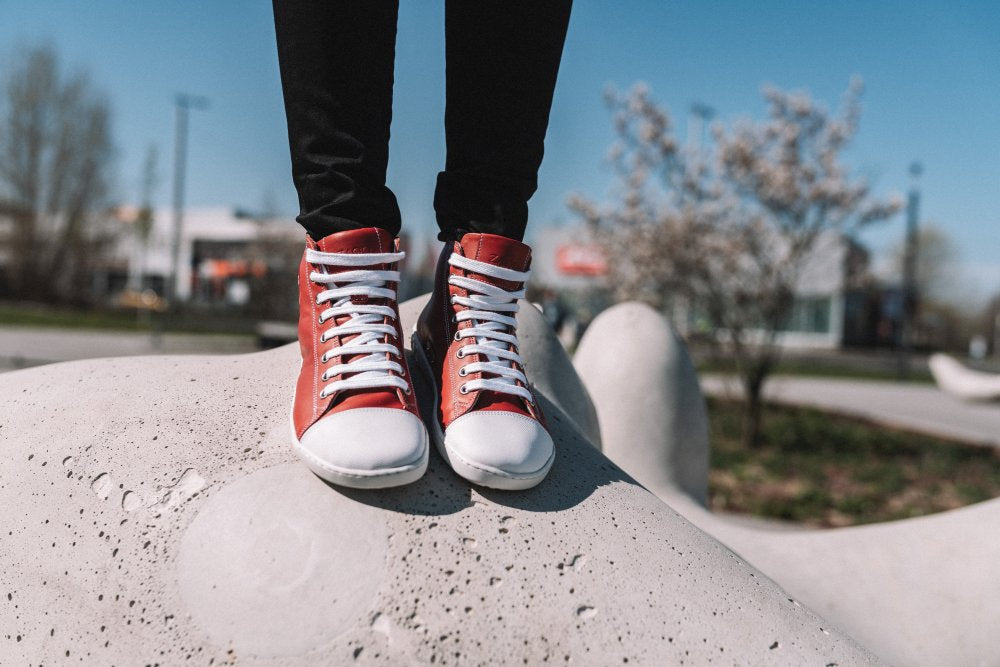 Eine Person steht auf einer großen, glatten Betonkonstruktion und präsentiert die CHUQQS Red High-Top-Sneaker von ZAQQ, gefertigt aus hochwertigem Nappaleder. Im Hintergrund ist eine Parklandschaft mit Bäumen und Gebäuden unter einem klaren blauen Himmel zu sehen.