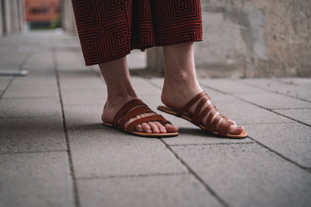 Eine Person trägt die FLAQE Brown Sandalen von ZAQQ und einen fließenden, gemusterten Rock und steht auf einem Betonweg. Im Hintergrund ist eine verschwommene Steinmauer zu sehen, die die Essenz urbaner Schlichtheit perfekt einfängt.