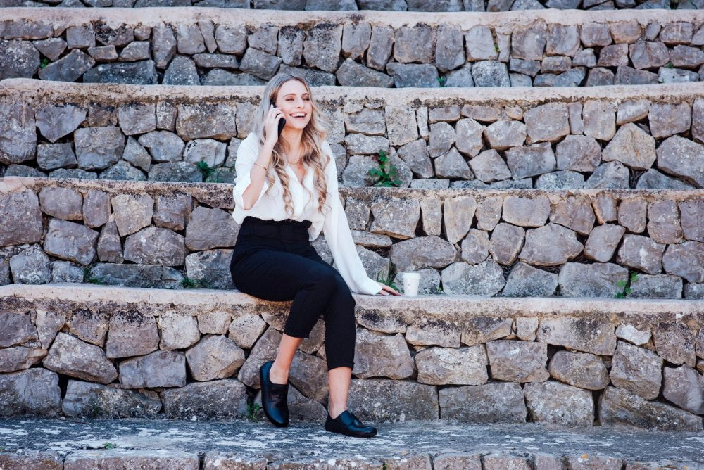 Eine Frau mit langen Haaren sitzt auf Steinstufen und ist tief in ihr Smartphone vertieft. Sie trägt ein elegantes Business-Outfit bestehend aus weißer Bluse und schwarzer Hose, ergänzt durch stylische ZAQQ-Schuhe, genauer gesagt das Modell IQON Black im Derby-Schnitt-Design. Neben ihr steht eine Tasse, alles vor dem Hintergrund von strukturiertem Outdoor-Stein.