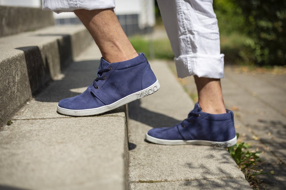 Eine Person in weißer Hose und blauen ZAQQ LANQASTER Navy Schuhen steigt draußen eine Betontreppe hinauf. Diese Schuhe aus Veloursleder sind seitlich mit einem Logo versehen und verkörpern den Charme eines sonnigen Tages.