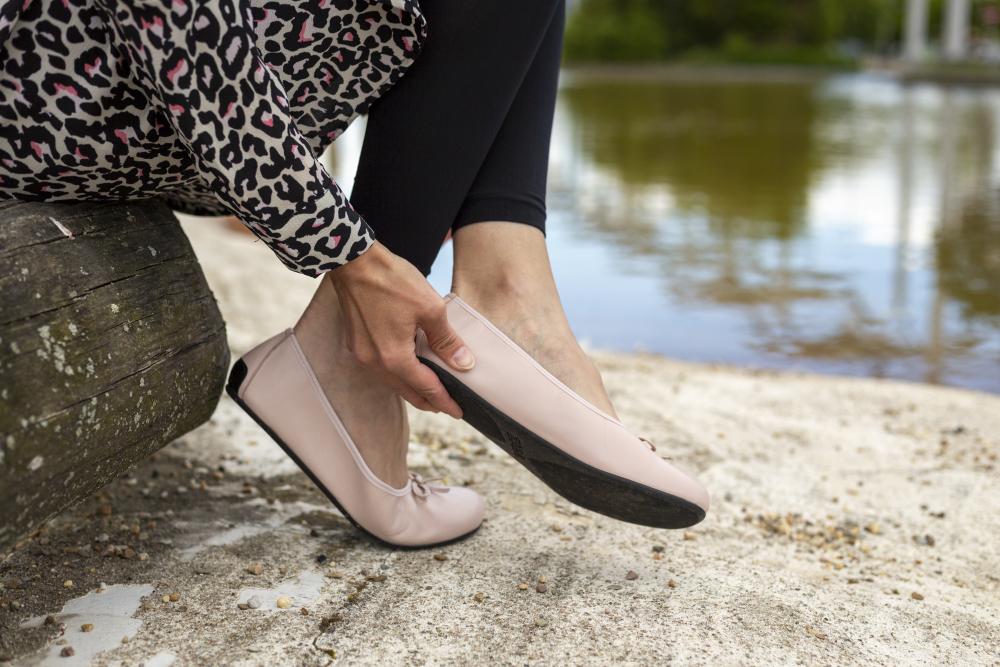 Eine Person sitzt auf einer Holzbank an einem Teich, elegant gekleidet in einem Mantel mit Leopardenmuster und schwarzen Leggings, und rückt ihre LOOQ Nude-Schuhe zurecht. Die Szene ist ruhig und heiter, mit Wasser und Bäumen im Hintergrund. Trotz des schicken Looks bleiben ihre ZAQQ-Schuhe bequem.