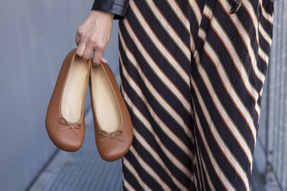 Eine Person hält ein Paar ZAQQ LOOQ Vegan Cognac Ballerinas mit Schleifen. Sie trägt gestreifte, schwarz-beige Hosen aus atmungsaktivem Material vor einer grauen Wand im Hintergrund.