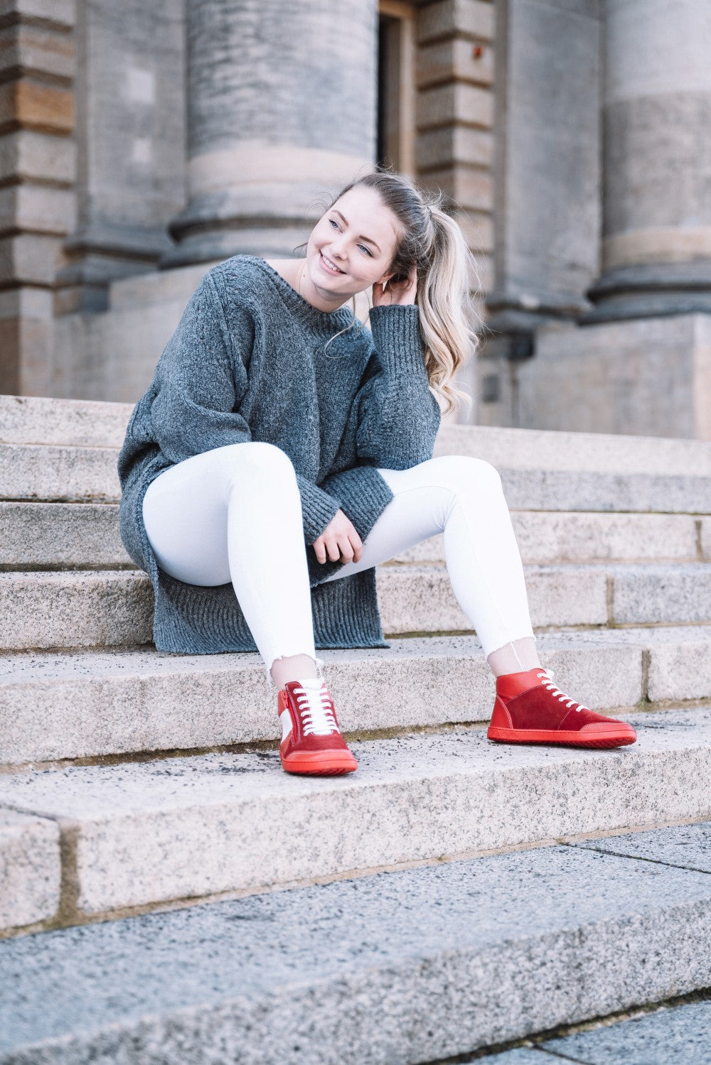 Eine Frau in grauem Pullover und weißer Hose sitzt lächelnd auf Steinstufen, ihre Haare sind zu einem Pferdeschwanz gebunden. Ihre LUQQ Red Sneakers von ZAQQ mit flexibler Kautschuksohle setzen vor dem Hintergrund eines Gebäudes mit großen Säulen einen Farbtupfer.