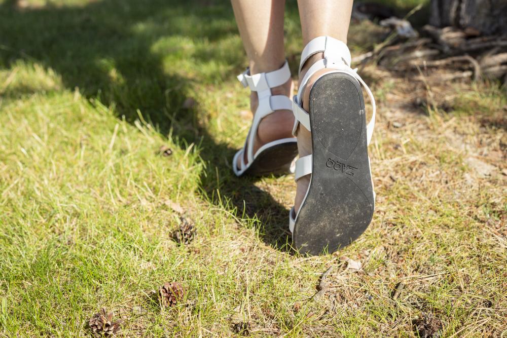 Eine Person schlendert in weißen ZAQQ MONIQ-Sandalen durch das Gras. Die Aufmerksamkeit wird auf die Sohle mit BOC-Aufdruck gelenkt. Das Sonnenlicht wirft spielerisch Schatten und betont die Textur des Grases und der kleinen, verstreuten Tannenzapfen.