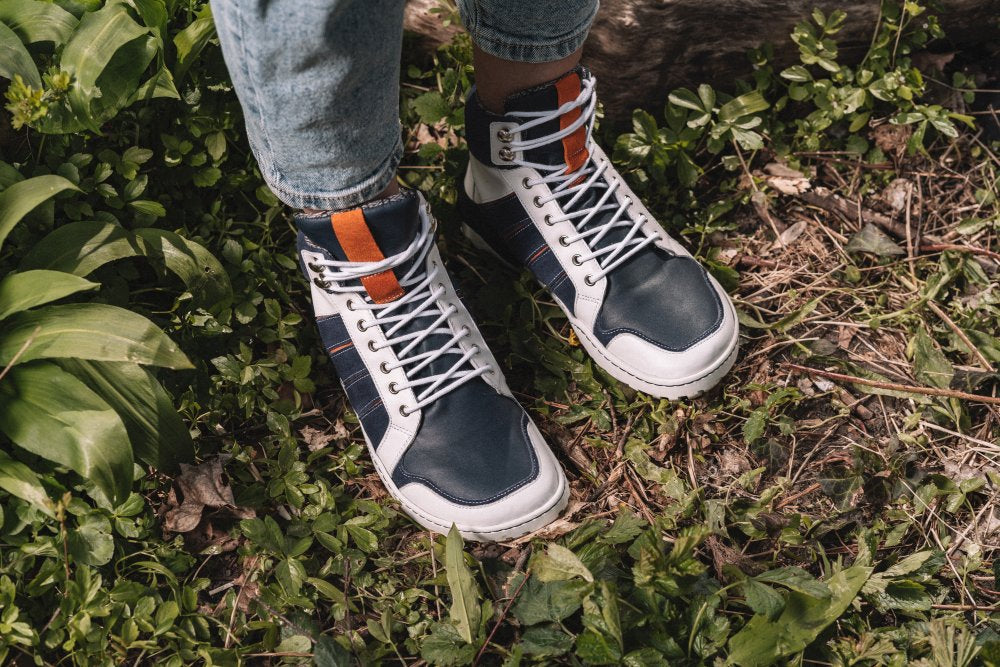 Nahaufnahme einer Person, die die ZAQQ MONTBLANQ Blue White Waterproof Sneaker mit orangefarbenen Akzenten und einer TERRA-Sohle trägt und auf grünem Gras inmitten kleiner Pflanzen steht. Das Outdoor-Schuhdesign ergänzt hellblaue Jeans perfekt und bietet eine ideale Mischung aus Stil und Funktionalität.