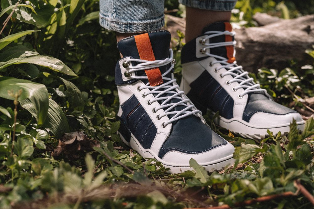 Eine Nahaufnahme zeigt die Füße einer Person, die in den wasserdichten High-Top-Sneakers MONTBLANQ Blue White von ZAQQ steckt, mit marineblauen, weißen und orangefarbenen Akzenten aus Nappaleder. Die Schuhe sind mit weißen Schnürsenkeln geschnürt und ruhen auf Gras inmitten üppigen Grüns. Sie eignen sich ideal als Outdoor-Schuh und passen gut zu hochgekrempelten Jeans.