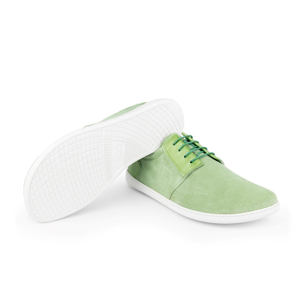 Ein Paar ZAQQ PIQUANT Lime Freizeitschuhe aus Nappa- und Veloursleder mit weißer Sohle. Ein Barfuß-Sneaker liegt auf der Seite, um die strukturierte Sohle hervorzuheben, während der andere aufrecht steht. Das minimalistische Design wird durch grüne Schnürsenkel aufgewertet.