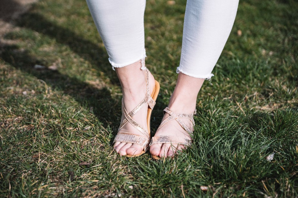 Eine Person in weißer Hose und taupefarbenen QEE Sandalen von ZAQQ, mit modernem Design, steht auf grünem Gras. Das filigrane Design der Barfuß-Sandale zeichnet sich durch eine flexible Sohle aus, während das Gras an einigen Stellen leicht trocken ist.