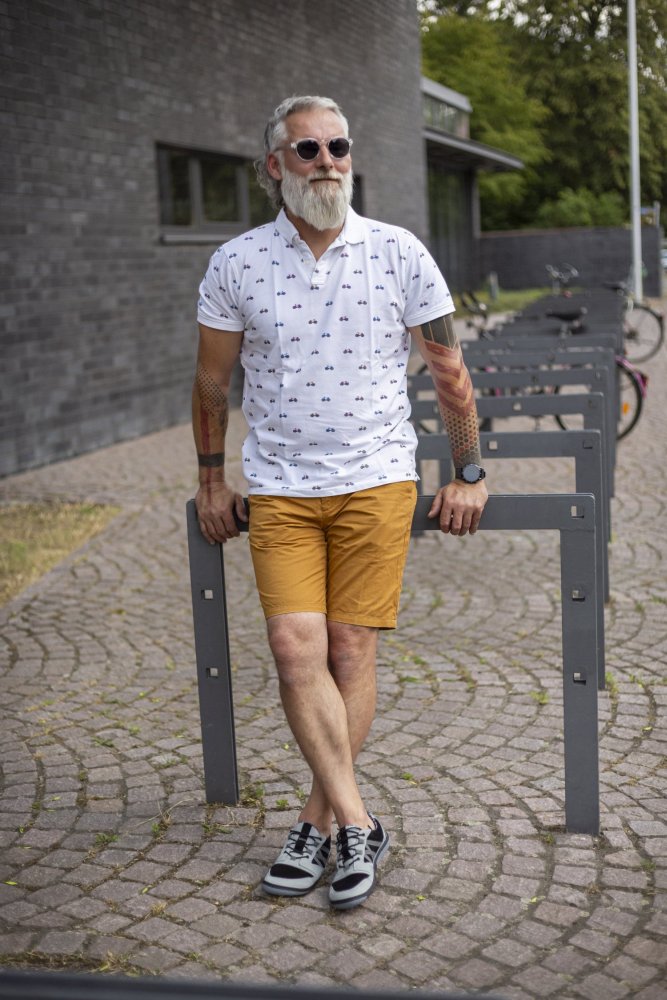 Ein Mann mit grauem Haar und Bart, Sonnenbrille, weiß gemustertem Hemd und senffarbenen Shorts lehnt an einem Fahrradständer aus Metall. Tattoos zieren seine Arme, während er in modischen schwarz-grauen ZAQQ QENDRIQ-Sneakers auf dem Kopfsteinpflasterweg steht.