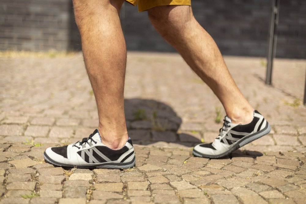 Eine Person in modischen schwarz-grauen ZAQQ QENDRIQ-Sneakern und braunen Shorts geht anmutig auf einem Kopfsteinpflasterweg.