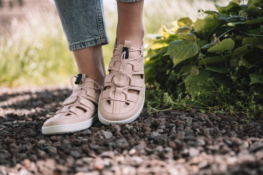 Eine Person, die den ZAQQ QLEAR Light Pink Sneaker mit Cut-Out-Design und schwarzen Akzenten trägt, der über hervorragenden Grip verfügt, steht auf einem Schotterweg. Im Hintergrund sind grüne Blattpflanzen zu sehen, die das Gefühl eines Outdoorschuh-Abenteuers verstärken.