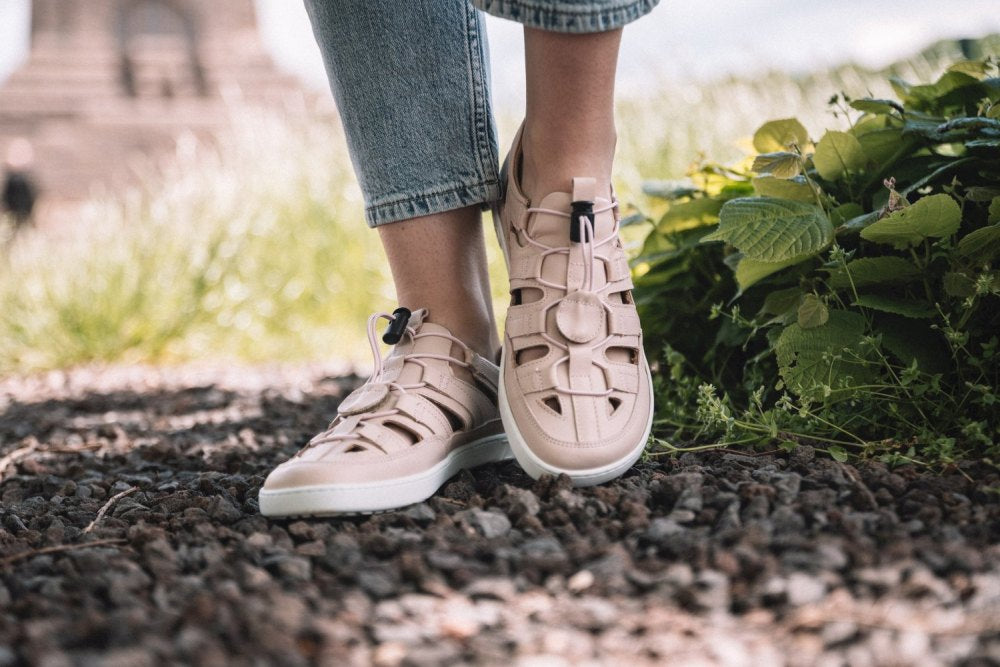 Eine Person, die QLEAR Light Pink-Sandalen von ZAQQ trägt, steht auf einem von Grün umgebenen Kiesweg. Diese Sandalen haben ein Gitterdesign und weiße Sohlen und bieten hervorragenden Halt für Outdoor-Abenteuer. Der Hintergrund ist verschwommen und suggeriert eine ruhige Naturkulisse.