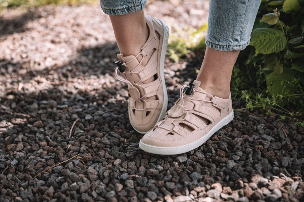 Eine Person in hellrosa QLEAR-Sandalen mit starkem Grip von ZAQQ steht auf einem Kiesweg. Sie trägt hellblaue Jeans und im Hintergrund sind ein paar grüne Pflanzen zu sehen, die für eine ruhige Außenatmosphäre sorgen.