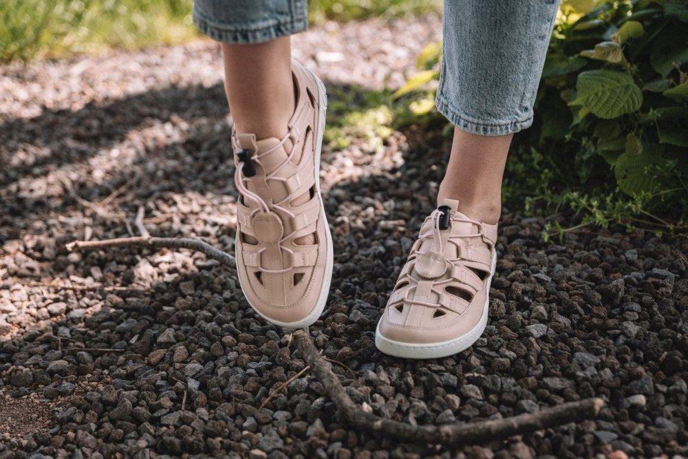 Nahaufnahme einer Person, die QLEAR Light Pink-Sandalen von ZAQQ mit weißen Sohlen trägt und selbstbewusst auf einem Kiesweg steht. Kombiniert mit hellblauen Jeans, die über den Knöcheln hochgekrempelt sind, bieten diese Sandalen beeindruckenden Halt. Im Hintergrund ist üppiges grünes Laub zu sehen.