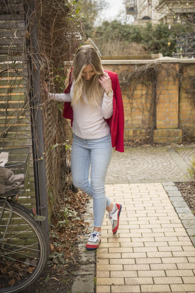 Eine Frau mit langen blonden Haaren schlendert einen Ziegelweg entlang, gekleidet in das weiße Oberteil QOLETTE von ZAQQ und hellblaue Jeans. Sie hängt ihre rote Jacke lässig über die Schulter und strahlt lässige Eleganz aus. In der Nähe lehnt ein Fahrrad an einer Mauer aus Ziegeln und Weinreben. Ihr mühelos schicker Stil verkörpert perfekt die ZAQQ Kollektion.

