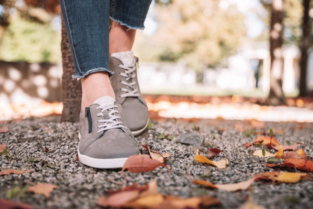 Eine Person, die ZAQQs QOMFY Wide Grey Sneakers und Blue Jeans trägt, steht auf einem mit Herbstlaub bedeckten Kiesweg. Im Hintergrund sind Bäume mit verschwommenem Laub zu sehen, die einen sonnigen Herbsttag suggerieren und bei jedem Schritt Behaglichkeit ausstrahlen.