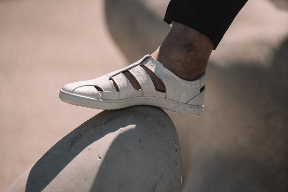 Eine Person, die die stylischen QRUISE White Sneaker von ZAQQ aus Nappaleder mit Cut-Out-Design trägt, stellt ihren Fuß lässig auf einen glatten Stein. Das moderne Aussehen und die Atmungsaktivität der Sneaker bilden einen schönen Kontrast zum verschwommenen, sonnenbeschienenen Hintergrund.