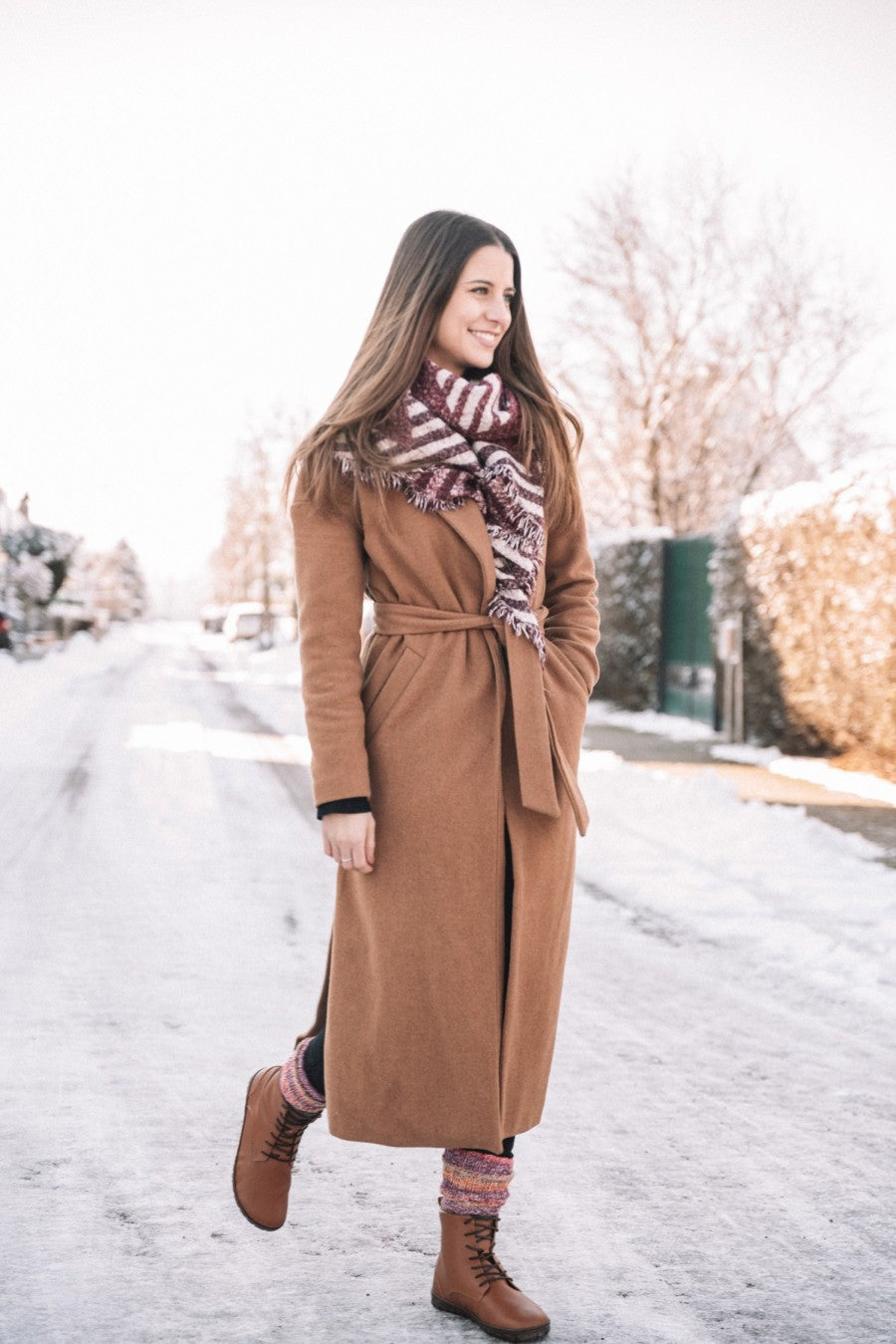 Eine Frau schlendert eine verschneite Straße entlang und trägt QUINTIC Winter Vegan Tabaco Stiefeletten von ZAQQ mit Kautschuksohle. Sie trägt einen langen braunen Mantel und einen gemusterten Schal, ihr langes Haar fällt herab, während sie lächelt und zur Seite blickt. Schneebedeckte Bäume und Häuser säumen die Straße unter einem hellen, klaren Himmel.