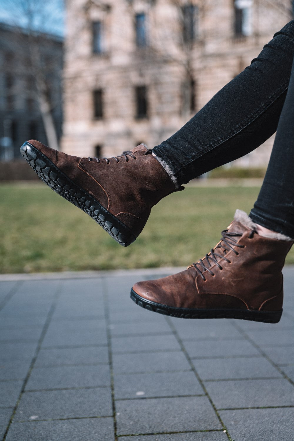 Eine Nahaufnahme zeigt eine Person mitten im Sprung. Sie trägt die QUINTIC Winter Velours Brown Waterproof-Stiefel von ZAQQ mit braunem Schnürdesign und schwarzen Sohlen und dazu dunkle Hosen. Im Hintergrund sind ein Steingebäude und ein Stück grünes Gras zu sehen, leicht verschwommen, um eine Stadtlandschaft an einem klaren Tag zu evozieren und das Gefühl von Bewegung und Freiheit zu verstärken.