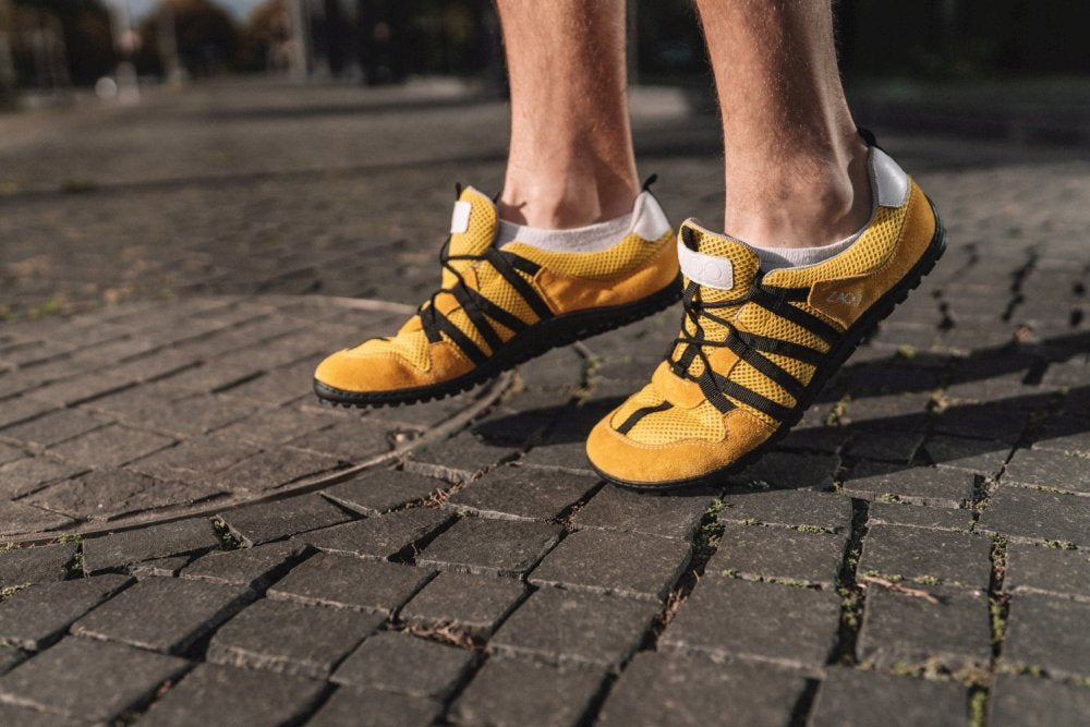 Nahaufnahme einer Person, die auf einem Kopfsteinpflasterweg läuft und die RIQ Yellow Sneaker von ZAQQ trägt. Diese Barfußschuhe bieten mit ihrem leuchtend gelben Design, akzentuiert durch schwarze Streifen und weiße Details, ein flexibles Tragegefühl. Der Fokus liegt auf den Schuhen und den Unterschenkeln.