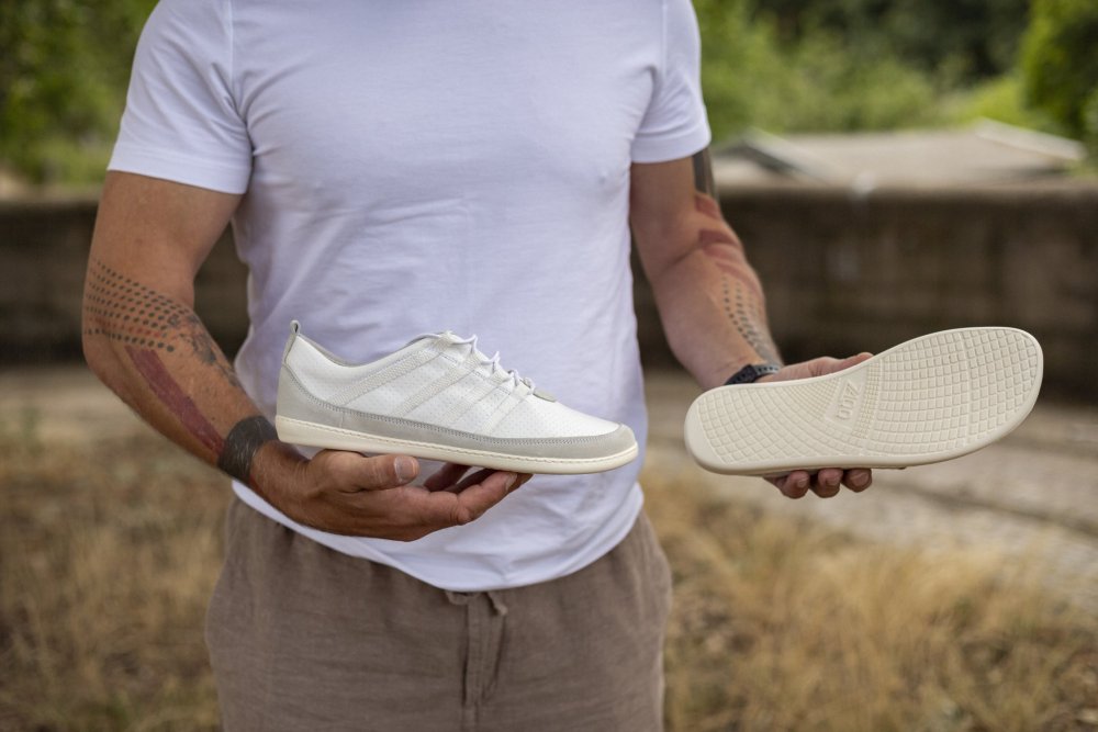 Eine Person in weißem Hemd und brauner Hose hält in einer Hand einen SPARQ Low White Sneaker von ZAQQ und präsentiert mit der anderen die Sohle. Im Hintergrund sind verschwommenes Grün und eine Steinmauer zu sehen, was den Komfort unterstreicht, der das Design dieses Barfußschuhs ergänzt.