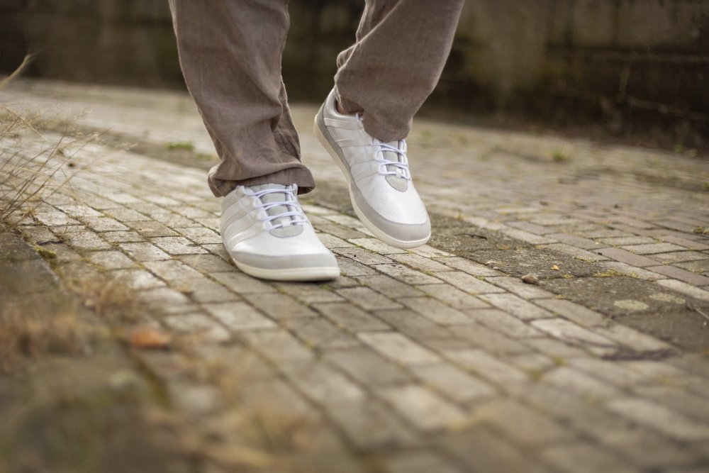 Eine Person in hellbraunen Hosen und den SPARQ Low White Sneakers von ZAQQ läuft auf einem Kopfsteinpflasterweg. Das Bild konzentriert sich auf den Unterkörper und zeigt die Sneakers in Bewegung. Gefallene Blätter und Gras zieren die Ränder des Weges und betonen den Tragekomfort bei jedem Schritt.