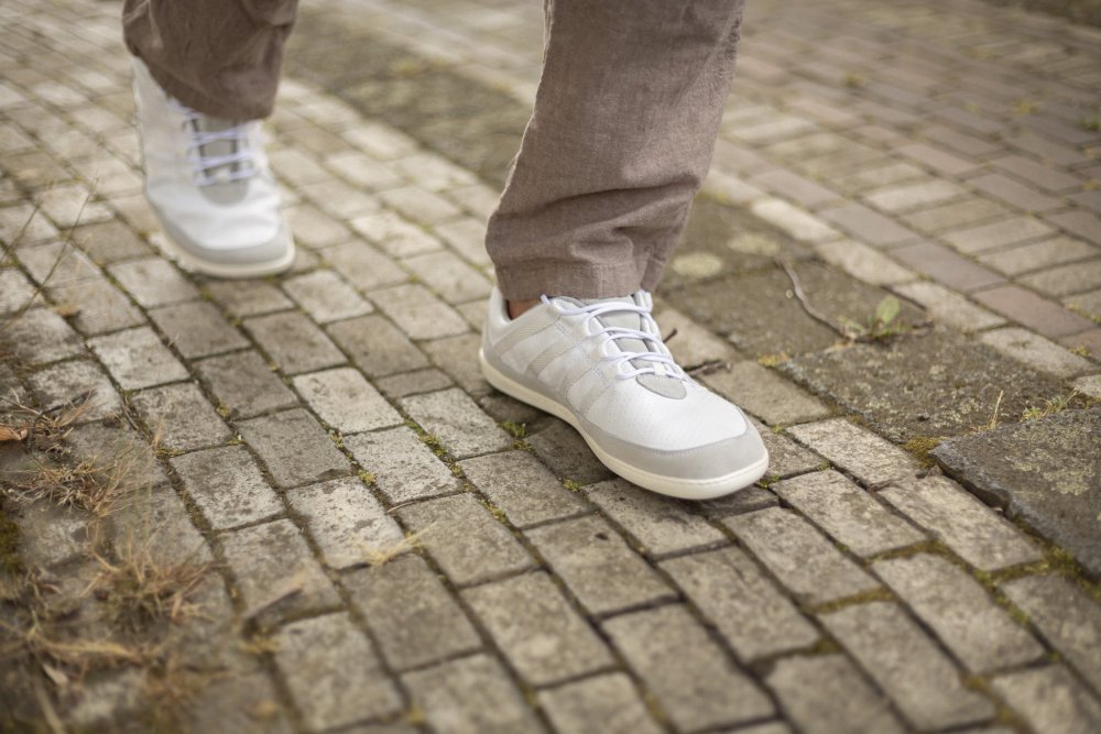 Nahaufnahme einer Person, die auf einem Kopfsteinpflasterweg läuft und ZAQQs SPARQ Low White-Sneaker und hellbraune Hosen trägt. Der Boden, der zwischen den Steinen mit Moos und Gras bedeckt ist, erhöht den Komfort bei jedem Schritt.