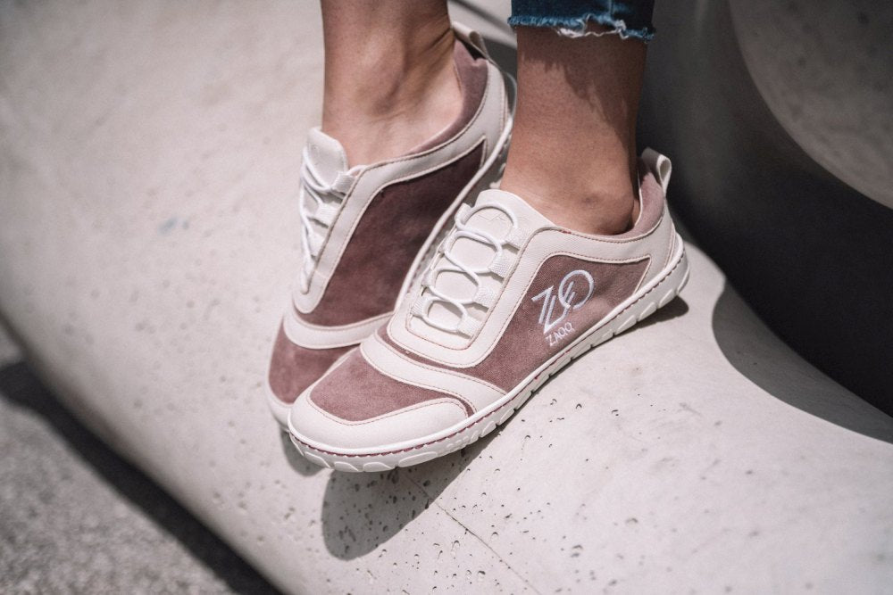 Nahaufnahme der Füße einer Person, die die stylischen SQILL Rose Sneaker von ZAQQ trägt, mit braunen und weißen Akzenten und dem Logo der Marke an der Seite. Diese Schuhe aus veganer Mikrofaser sind ideal, um auf einer großen, glatten Steinfläche zu stehen. Die Person trägt außerdem hochgekrempelte Jeans.