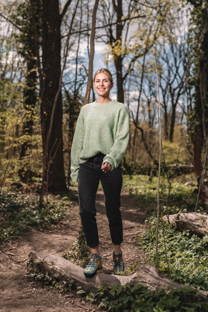 Eine Person in einem grünen Pullover und einer schwarzen Hose steht barfuß in ZAQQs SQOUT Blue Orange Waterproof-Schuhen auf einem Baumstamm im Wald, umgeben von Bäumen und Grün unter einem sonnigen Himmel. Das stylische Schuhwerk ist sowohl wasserdicht als auch atmungsaktiv und perfekt, um die Schönheit der Natur zu erkunden.