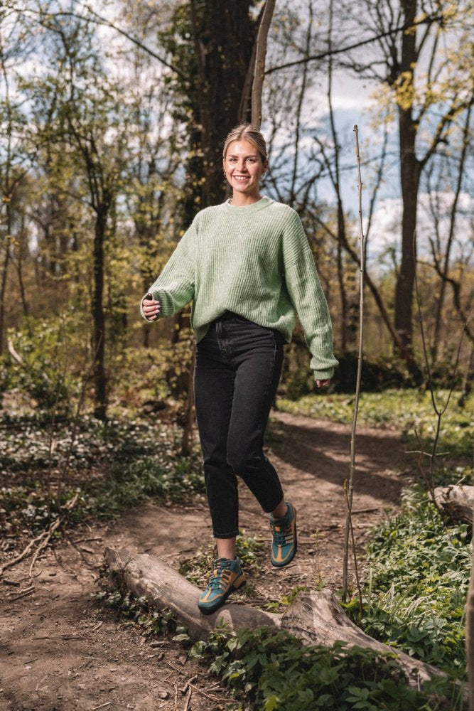 Eine Person in einem grünen Pullover und schwarzen Hosen geht lächelnd einen Waldweg entlang. Das Sonnenlicht fällt durch die Bäume und schafft eine warme und einladende Atmosphäre im üppigen Wald, während ihre wasserdichten Schuhe ZAQQ SQOUT Blue Orange sie bei jedem Schritt bequem halten.