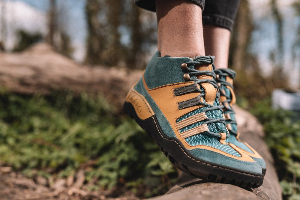 Nahaufnahme einer Person, die blaue und orangefarbene, wasserdichte SQOUT-Schuhe von ZAQQ trägt und auf einem Felsen steht. Der Hintergrund zeigt üppige Bäume und Grün, was eine belebende Umgebung im Freien suggeriert.