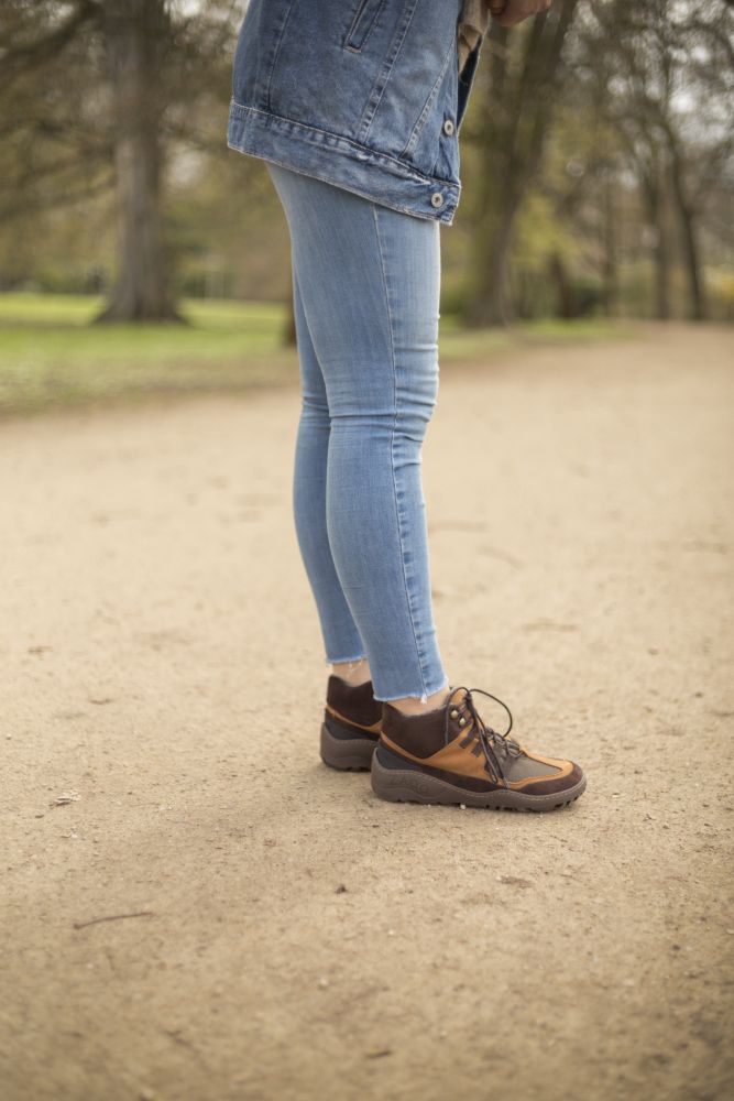 Eine Person in Blue Jeans, Jeansjacke und SQOUT Brown Waterproof Wanderschuhen von ZAQQ steht auf einem Sandweg in einem Park. Im Hintergrund sind Bäume und Gras zu sehen.