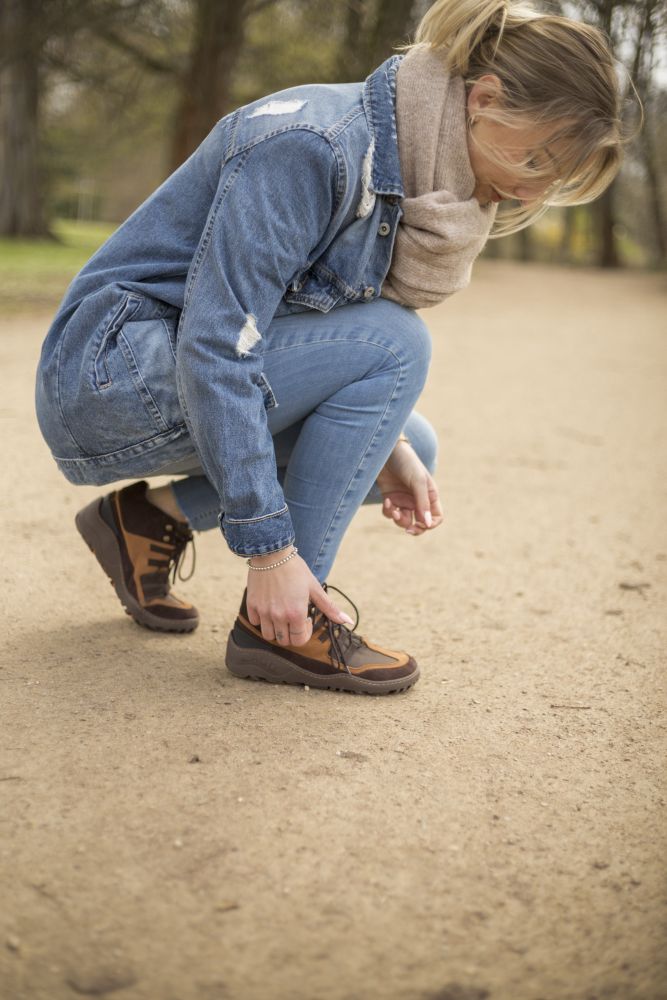 Eine Person in Jeansjacke und Jeans hockt auf einem Sandweg und schnürt die Schnürsenkel der SQOUT Brown Waterproof-Schuhe von ZAQQ. Im Hintergrund sind verschwommene Bäume und Grün zu sehen, die auf ein Abenteuer hinweisen, das jenseits des ausgetretenen Pfades wartet.