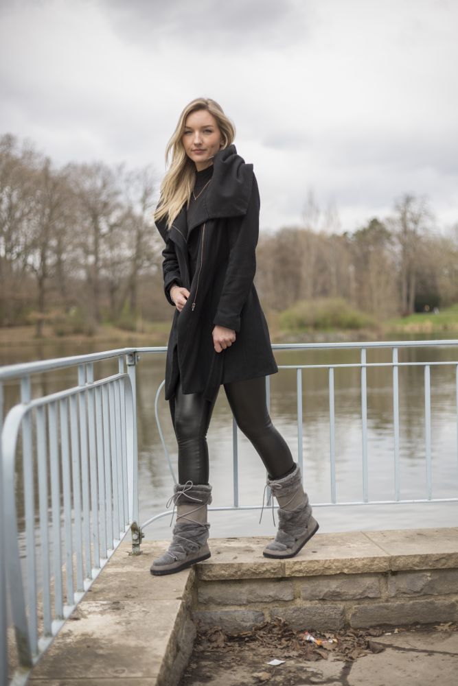 Auf einer Steinplattform an einem See stehend genießt eine Person mit langen Haaren das Barfußgefühl in TORQ Winterstiefeln von ZAQQ. Bekleidet ist sie mit einem langen schwarzen Mantel und dunklen Leggings. Die ruhige Szene wird durch kahle Bäume und einen bedeckten Himmel im Hintergrund ergänzt.